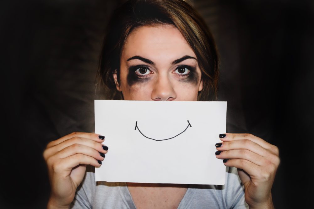 Woman with mascara smuged tears holding up a white paper with a smile on it.