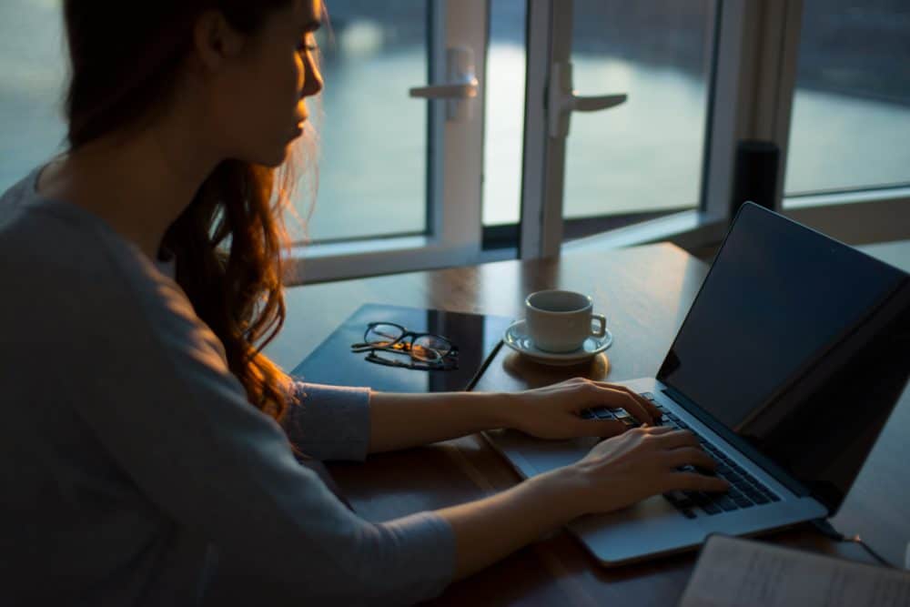 Woman sitting at a table on a laptop with a pair of glasses and cup of coffee next to her.