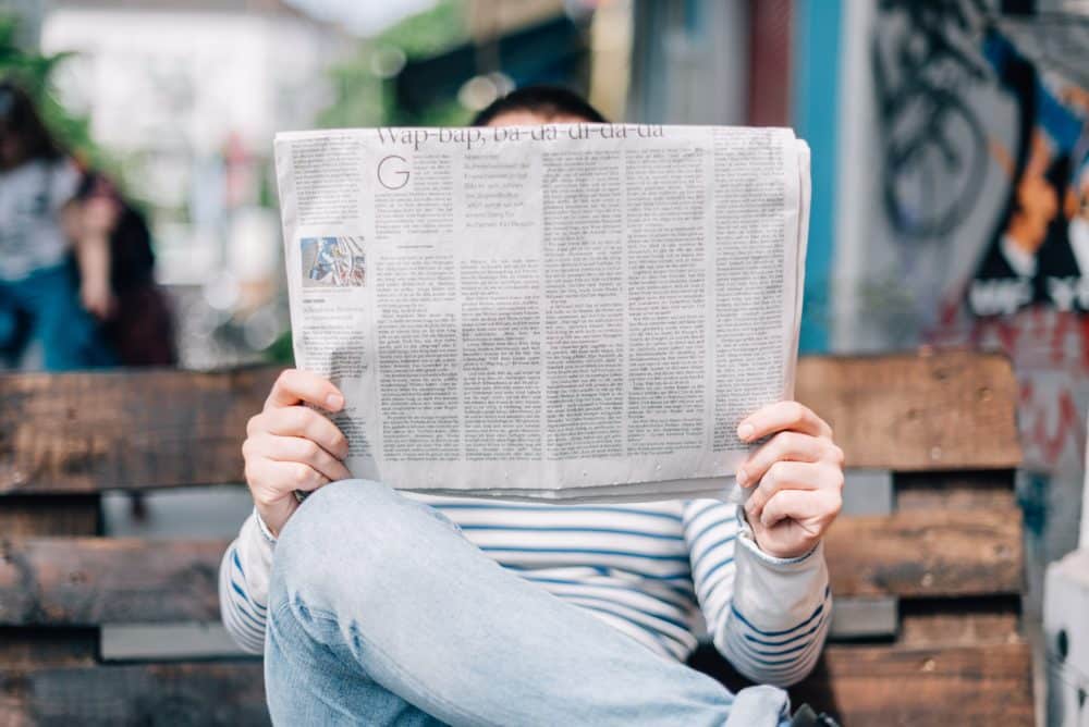 Person sitting on bench reading newspaper covering their face.