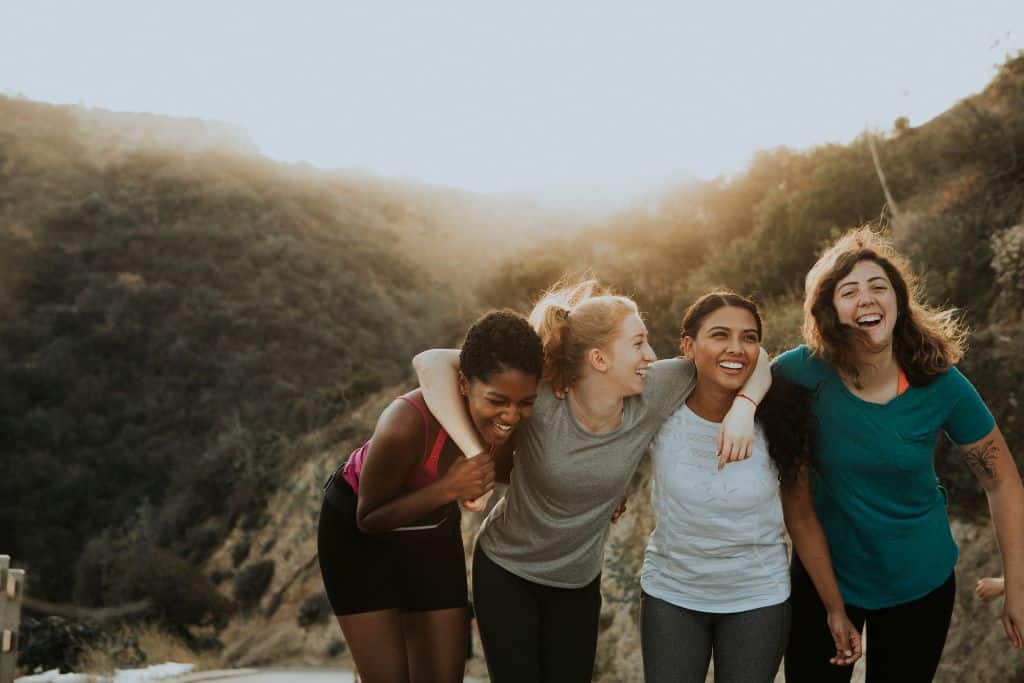 4 women link their arms and laugh while on a hike.