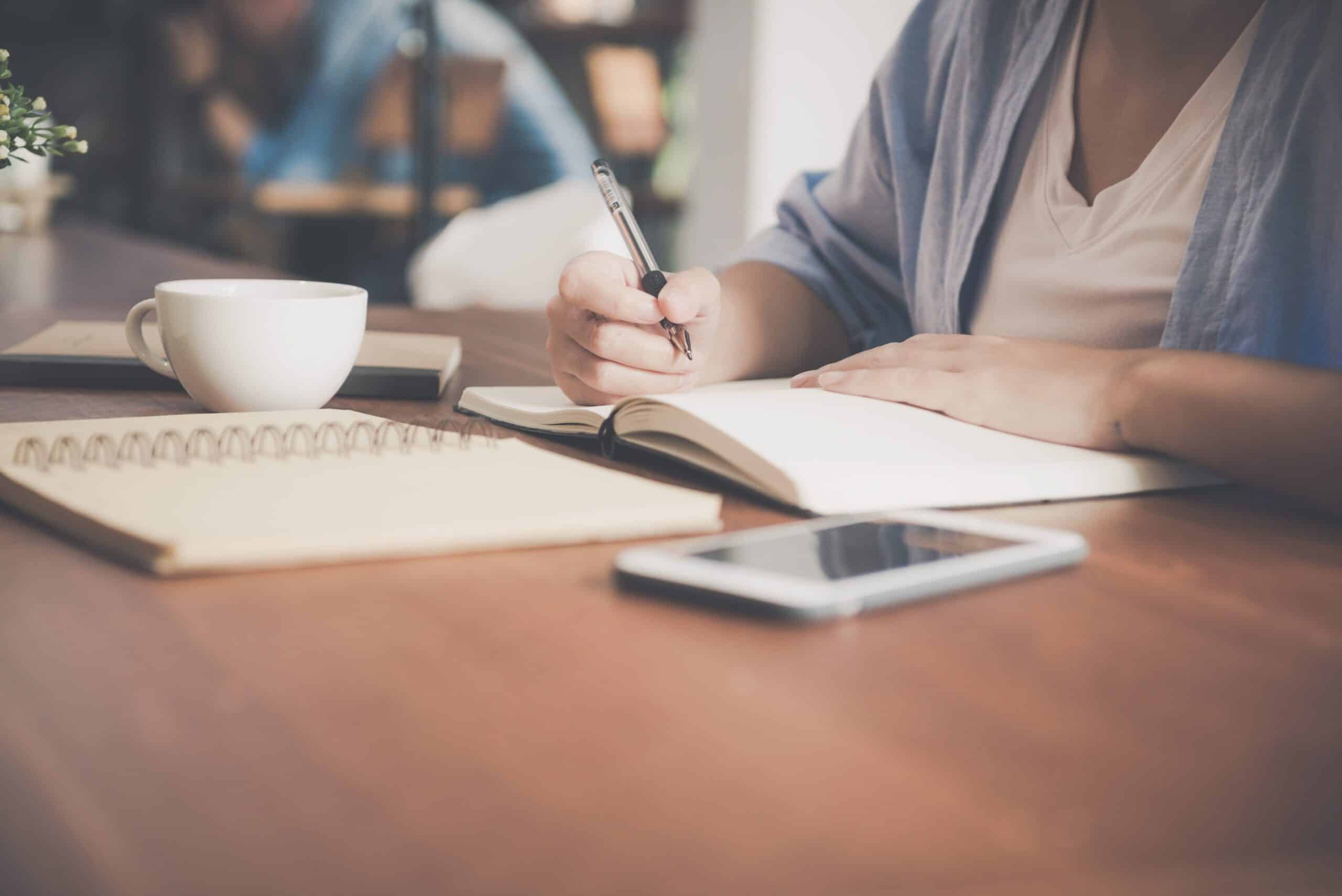 Woman writing in notebook with coffee and phone on table.