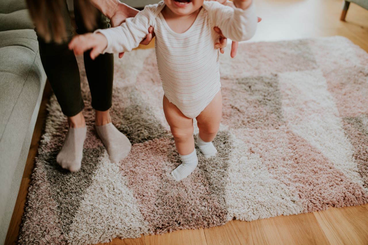 Woman in socks helping baby walk on carpet.