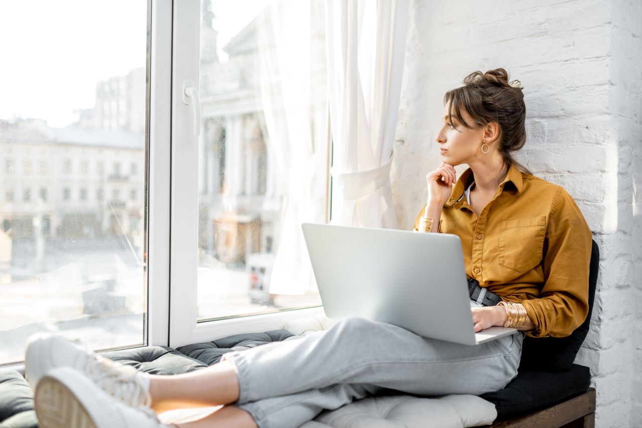Young woman dressed casually working on laptop while sitting on the window sill and looking out the window.