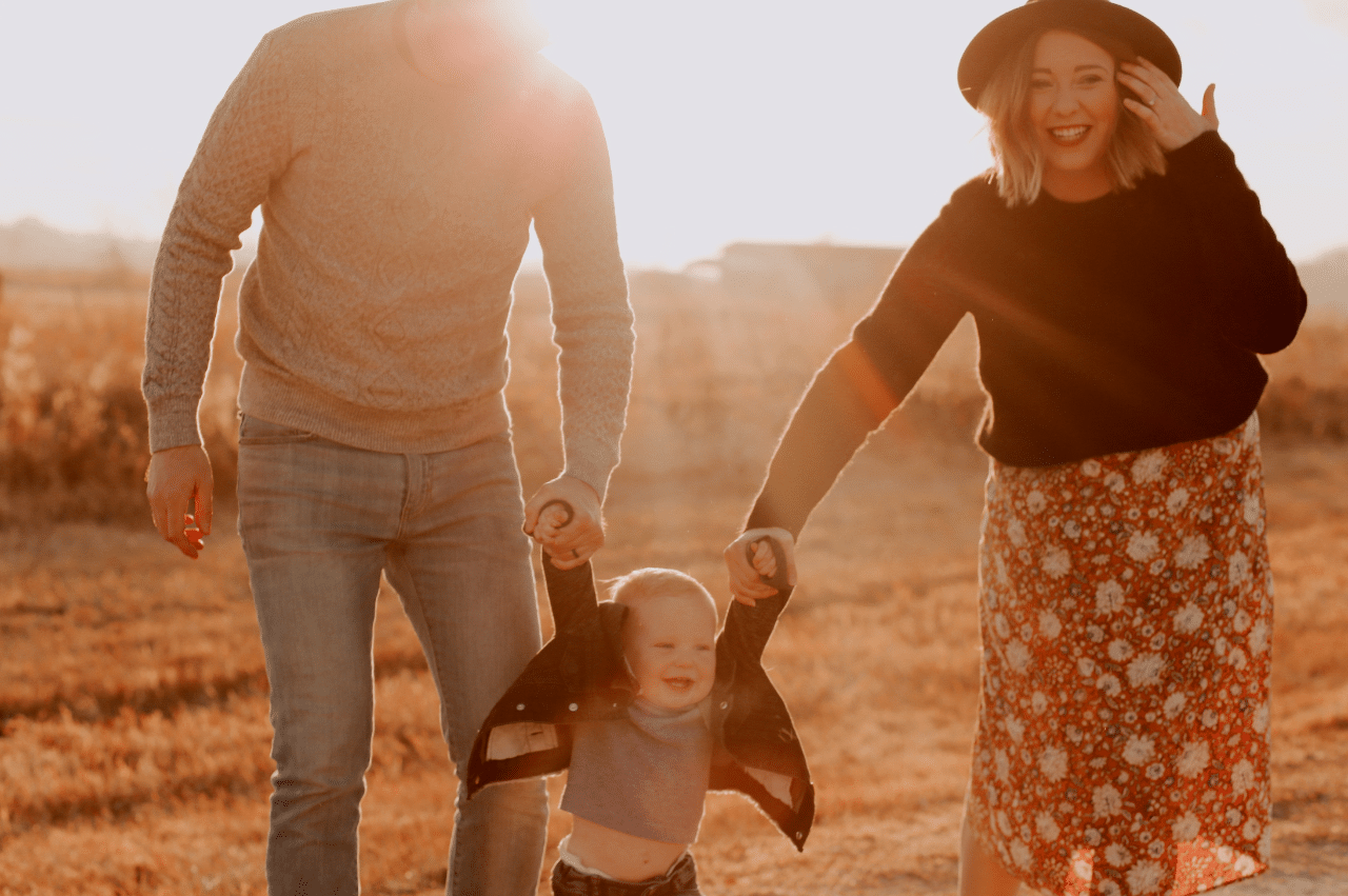 Father and smiling mother holding young child's hands as they walk toward the camera with the sun shining behind them.