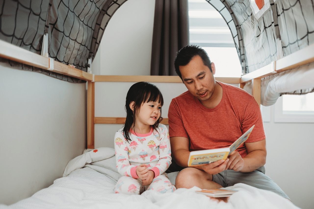 Man reading to a young girl while sitting on a bed.