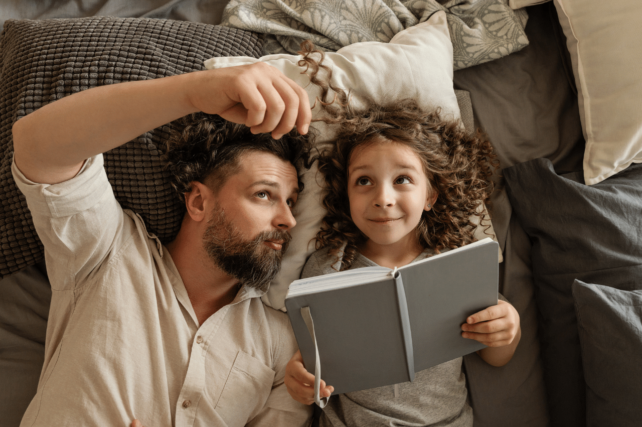 Dad and daughter laying on bed and looking up while reading a book.