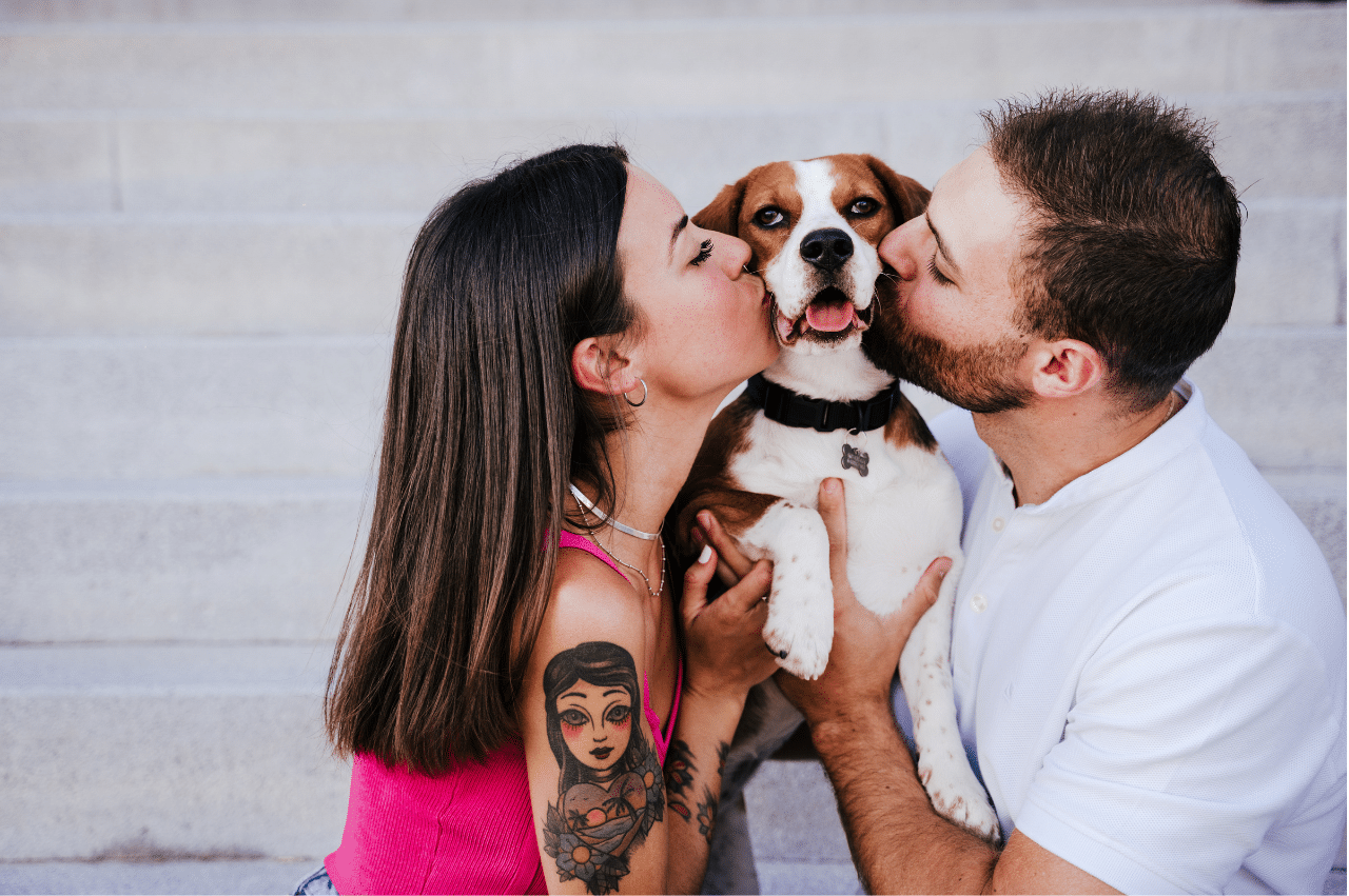 a white heterosexual couple holding up a medium size dog and kissing either side of its face
