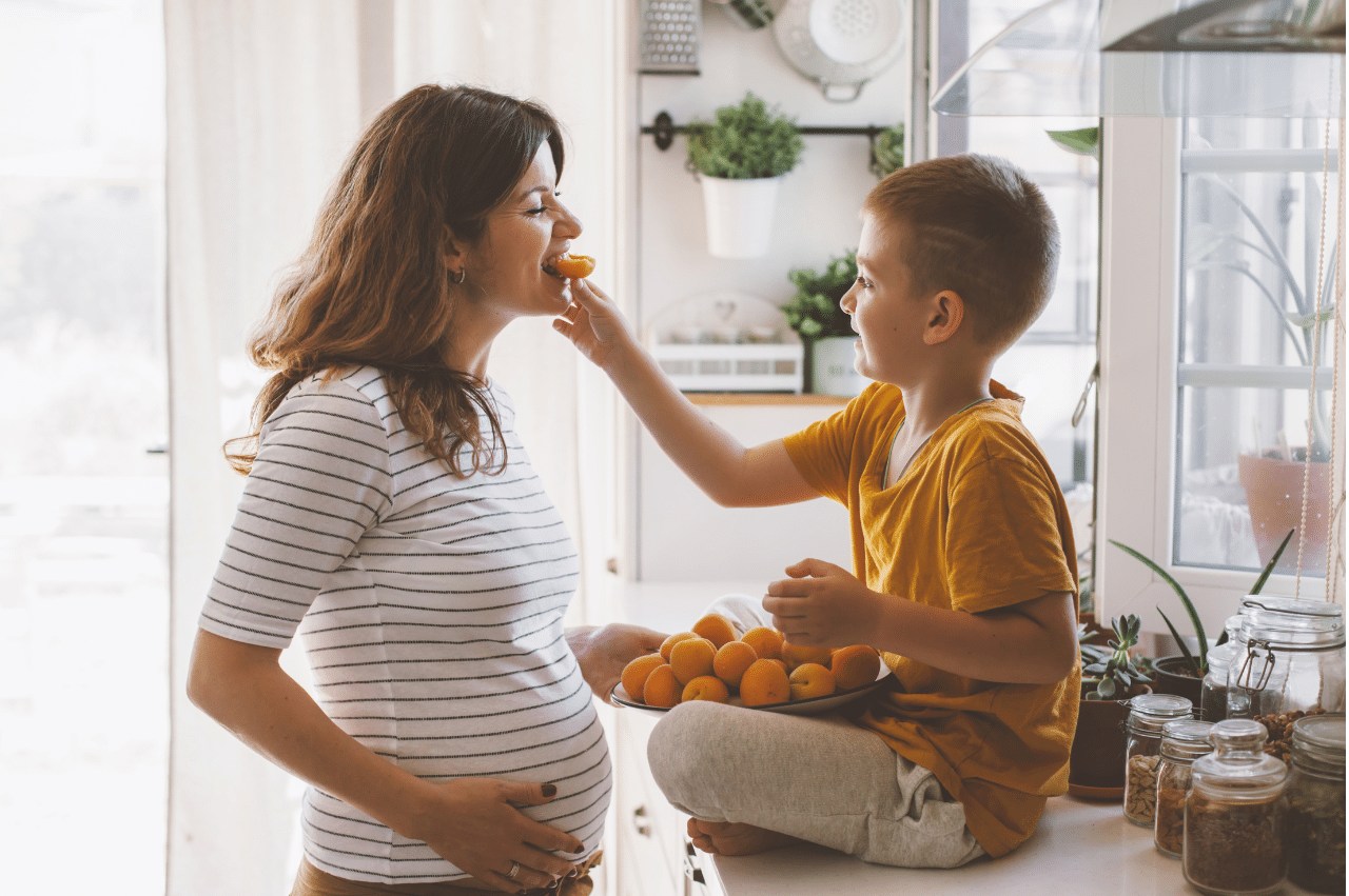 a little boy sitting on the kitchen counter feeding orange slices to his pregnant mom