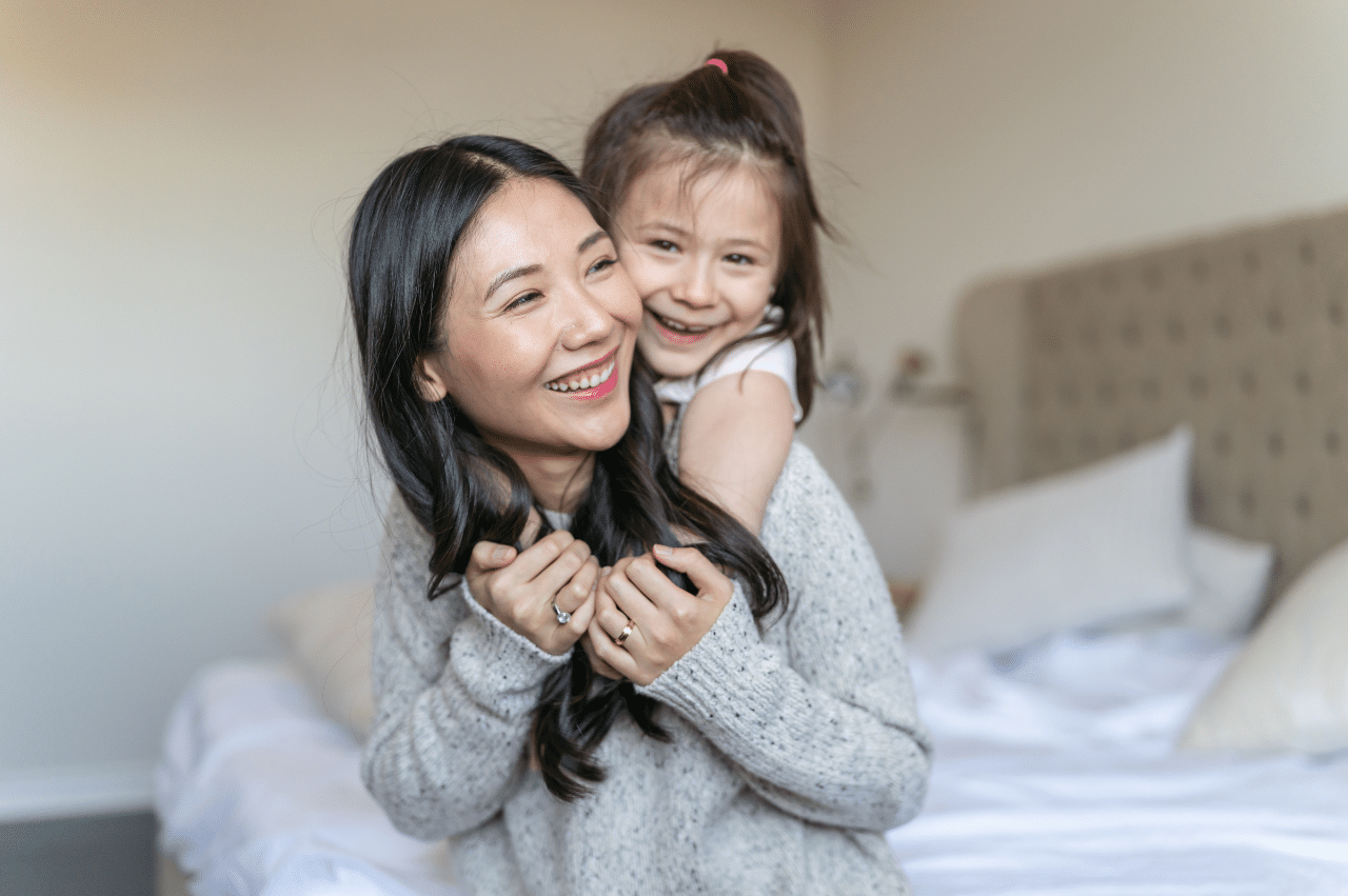 a woman sitting on a bed with her young daughter hugging her from over the shoulders