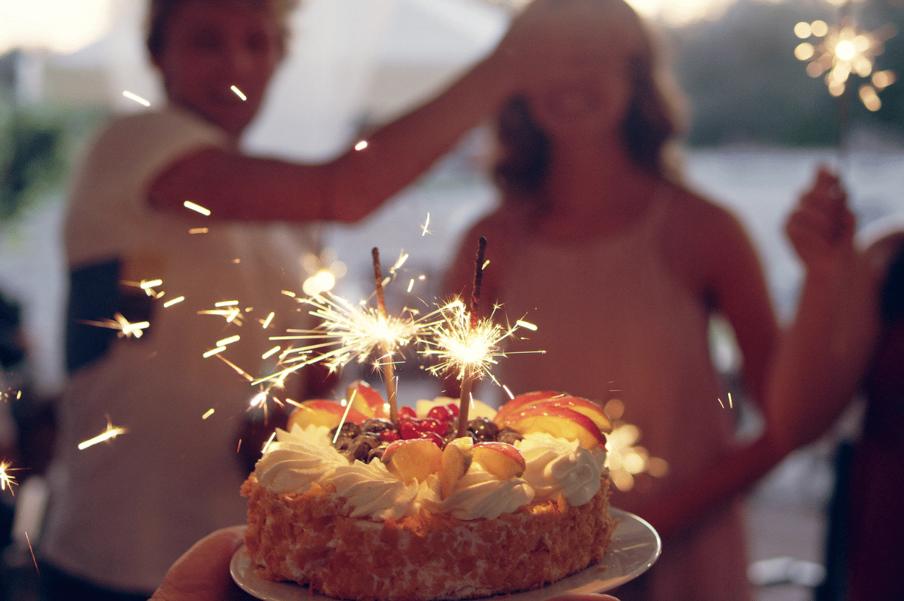 a birthday cake lit with sparklers in the foreground with a woman waiting in the background while someone else covers her eyes