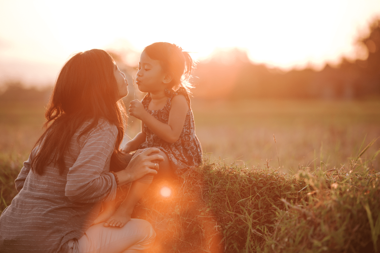 a backlit photo of a mother kissing her toddler-aged daughter in an open field