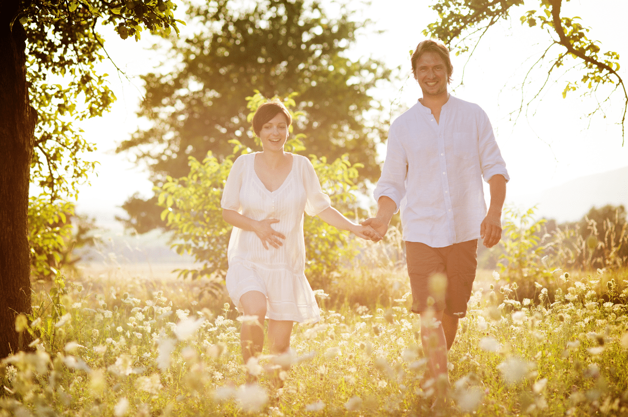 a young pregnant couple walking hand in hand through a meadow