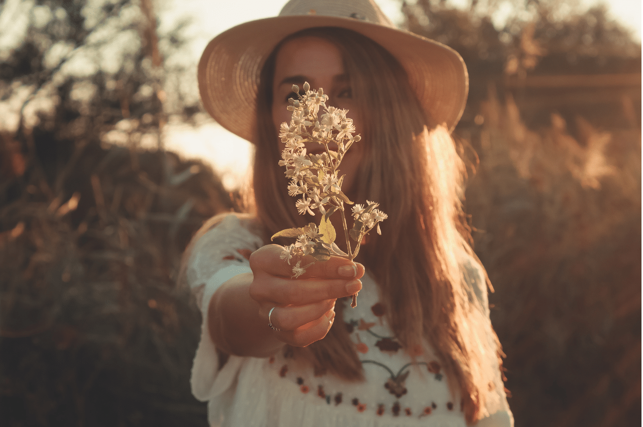 A woman in a straw hat holding out a sprig of white flowers that cover her face. She is in the center of the frame