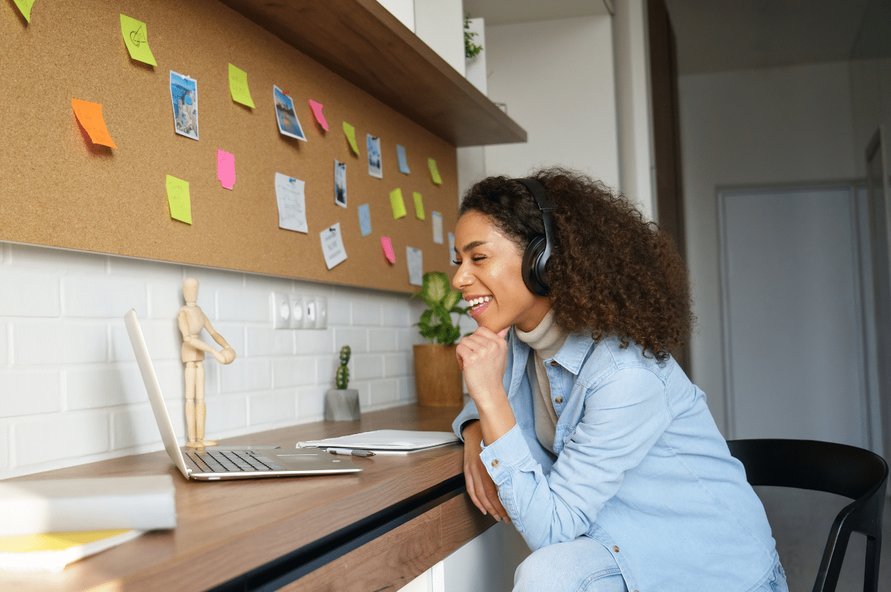 Woman sitting at a table with headphones on smiling at a laptop. There are sticky notes on a cork board above the table.