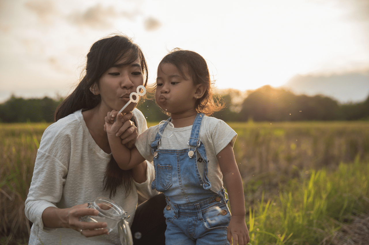 A woman and toddler blow through a bubble wand in a field