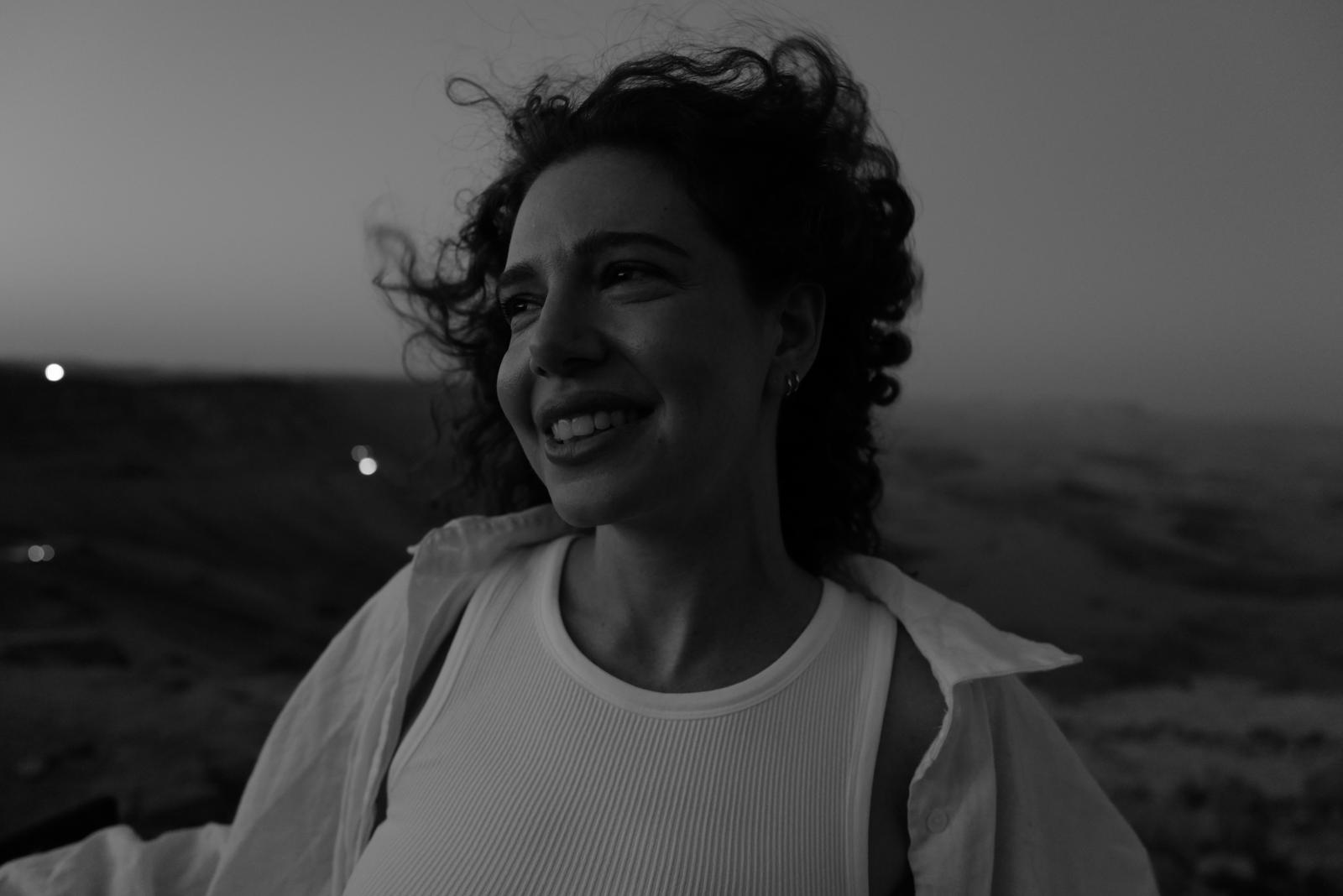 Black and white headshot of a woman smiling. Her hair is being blown in the wind and she is looking off to her right.