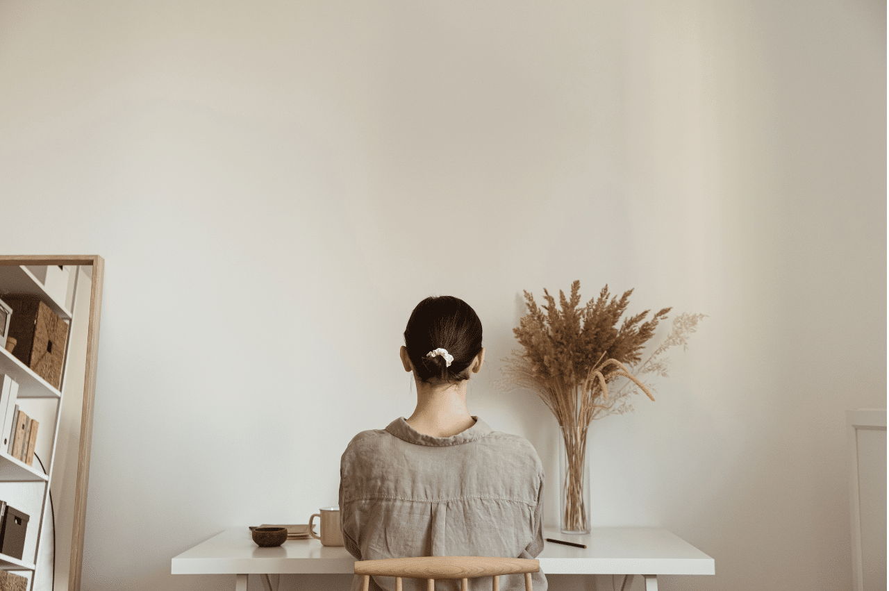 Back view of someone sitting at a desk. Their hair is in a bun and there is a mirror to the left of the desk. The colors of the room and furniture are neutral.
