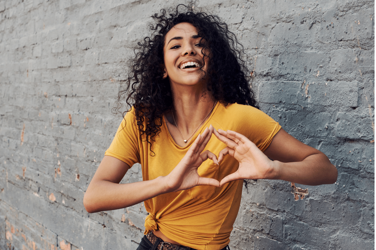 Woman smiling in front of a gray brick wall. She is wearing a yellow shirt and has her hands positioned to make a heart