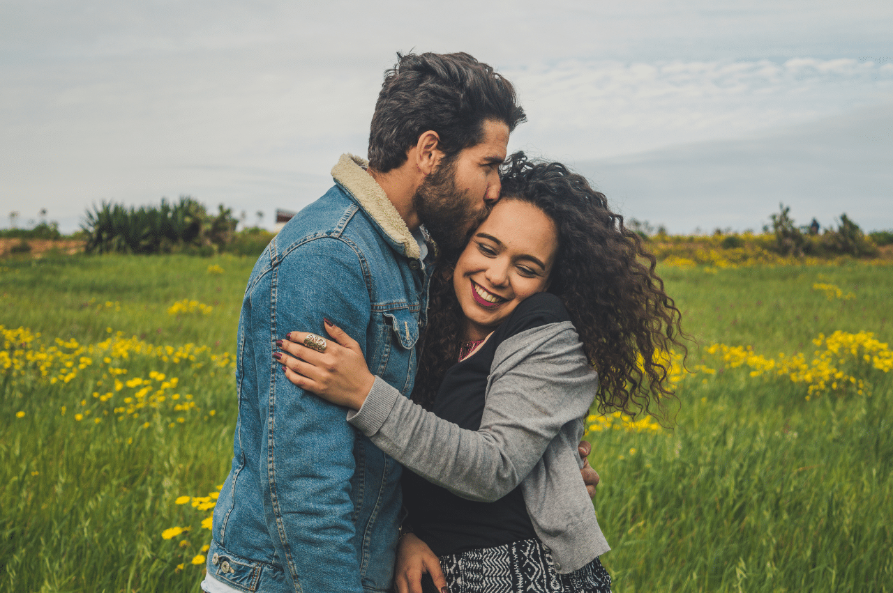 A couple is in a field of small yellow flowers. The person on the left is kissing the side of the person on the right's head.