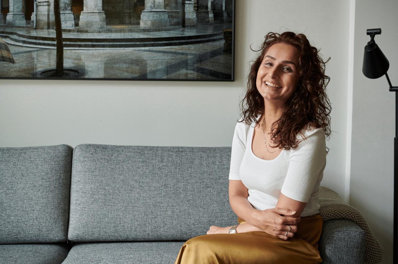 Woman sitting on a gray couch. Her arms are folded loosely and she is smiling.