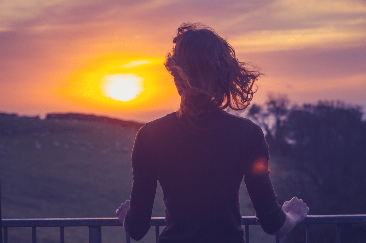 Waist up shot of a woman looking over a railing at a sunset/sunrise. The wind is moving her hair and she rests her hands on the railing.