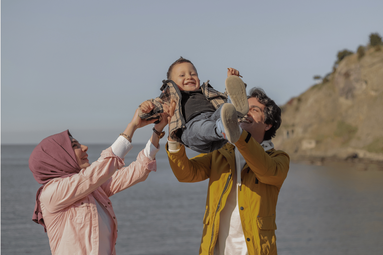 Parents swinging their toddler up in the air in front of a body of water. The child is laughing along with their parents.