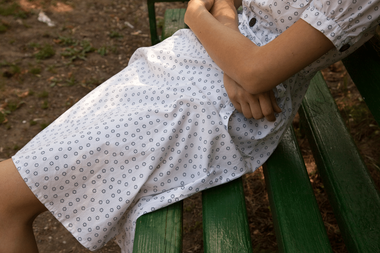 Abstract shot of a person sitting on a green bench with their arms crossed. They are wearing a white dress and the image shows from below the shoulders to above the knee.