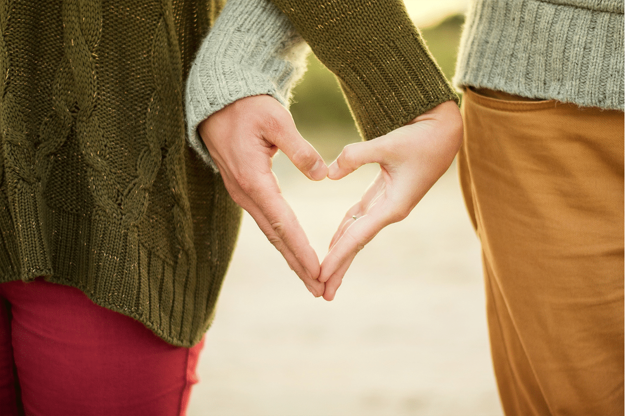 2 people putting their thumbs and fingertips to make the shape of a heart. They are wearing green knit sweaters and warm colored pants.