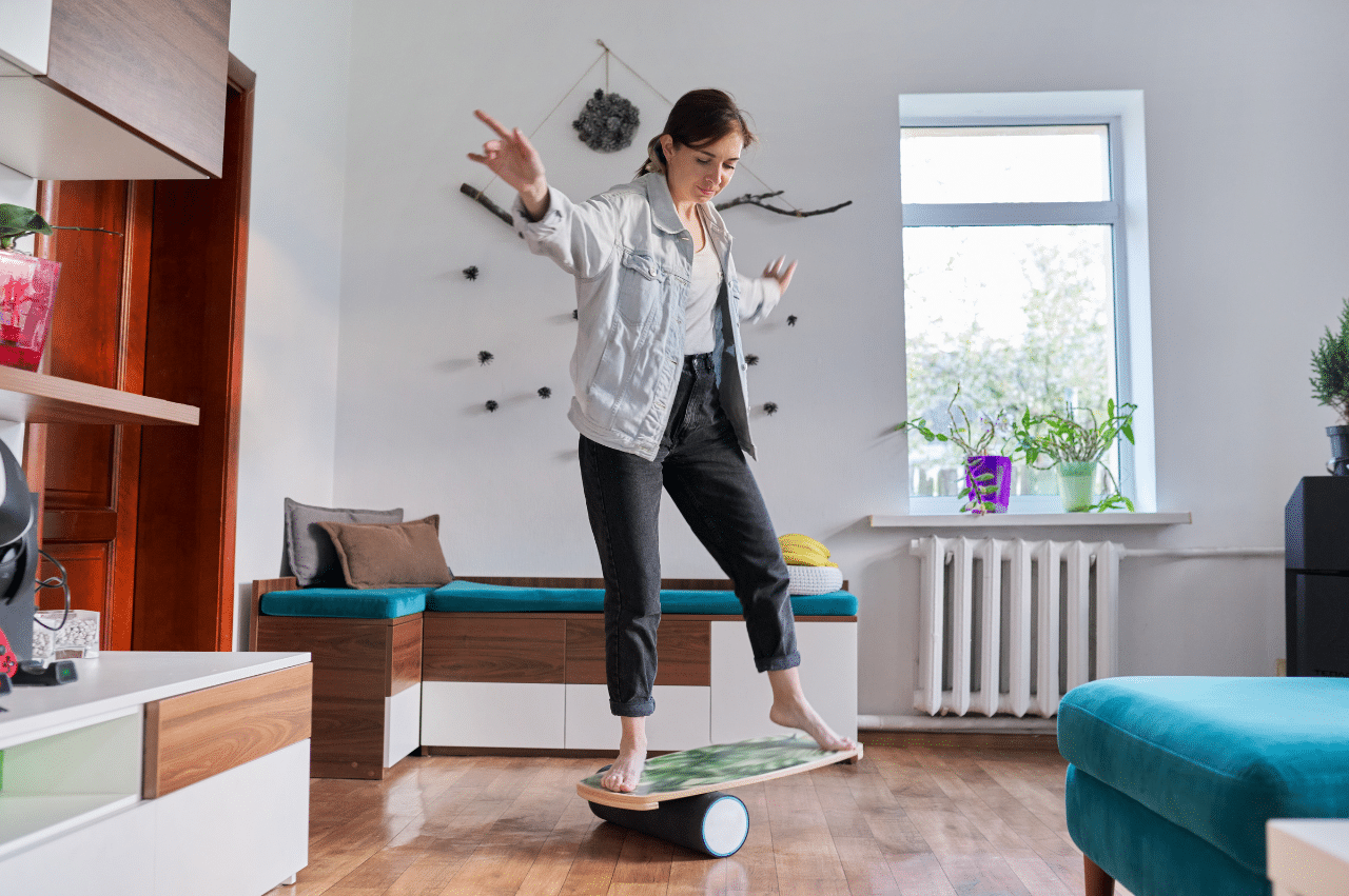 Woman balancing on a board on a rolling solid cylinder in a space that is minimally decorated and has white walls and light wood floors.
