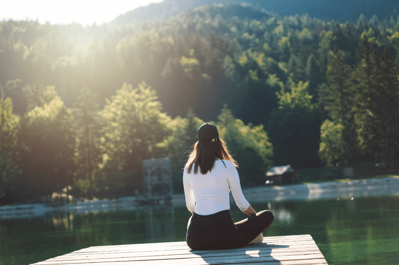 Woman sitting at the end of a dock facing a lake with a forest on the other side.