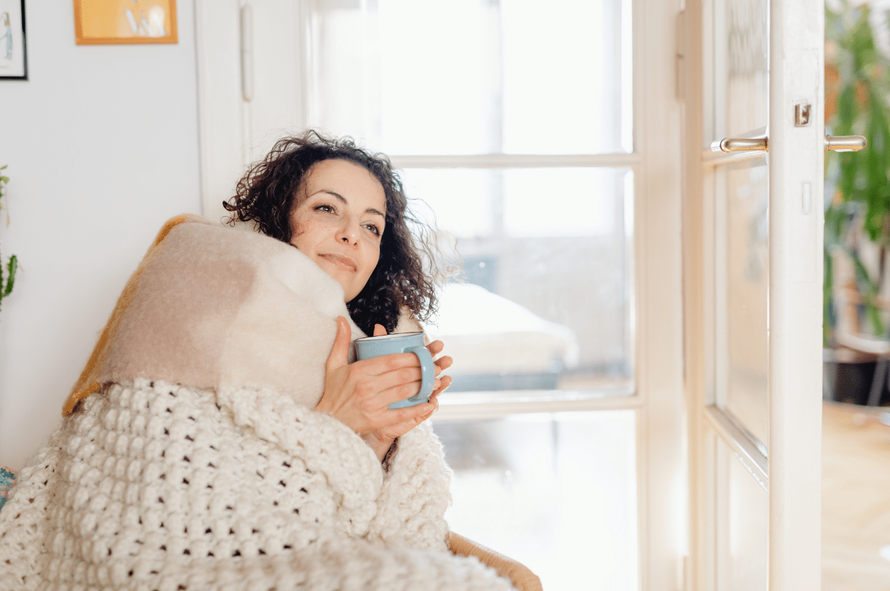 Woman in an oversized cream colored knit blanket holding a mug in a room with a big window with lots of natural sunlight