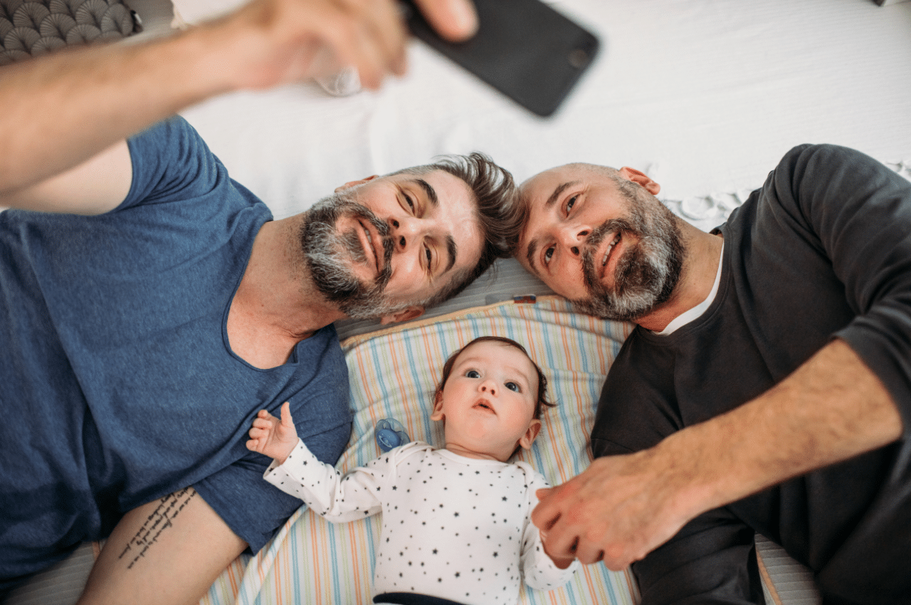 Photo of 2 dads laying with their baby on a bed