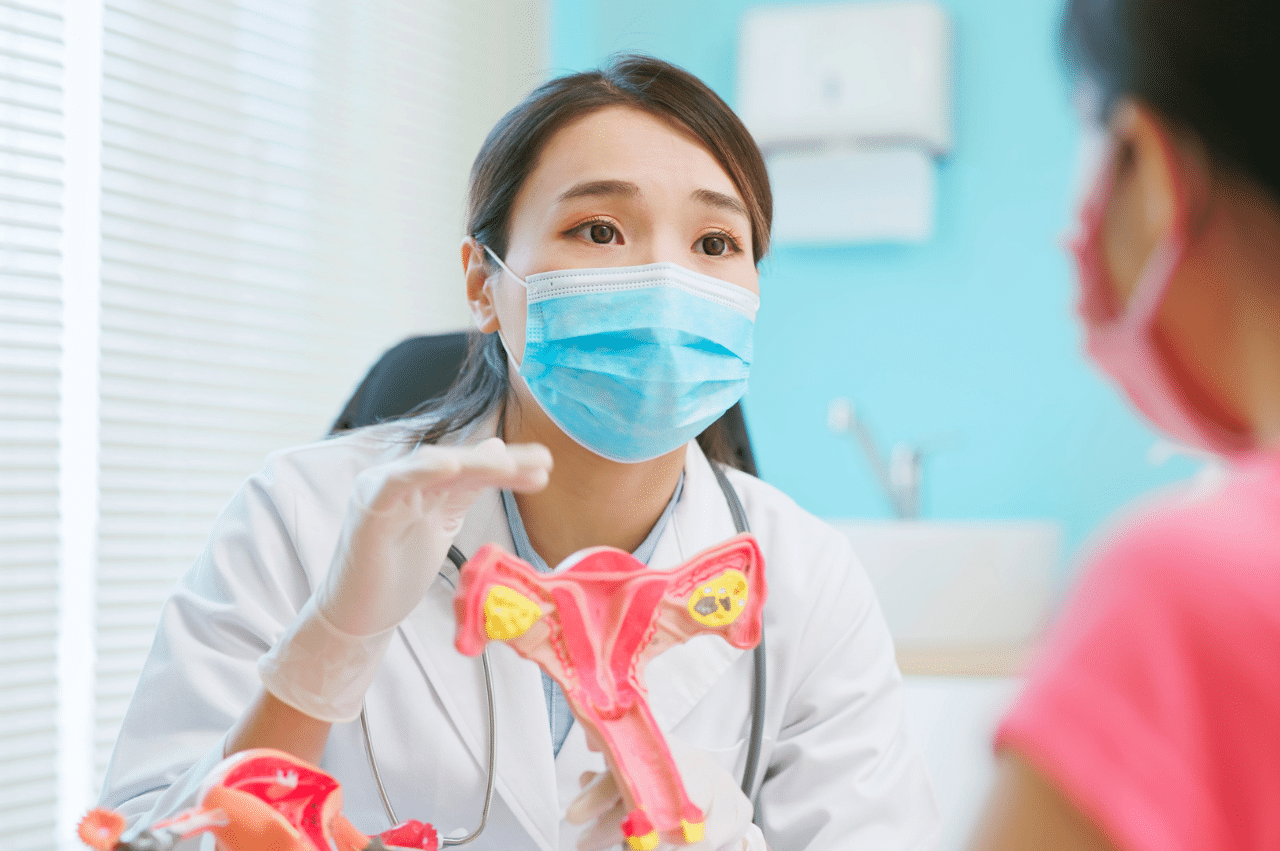 Photo of a doctor with a facemask on showing a model of a uterus to a patient