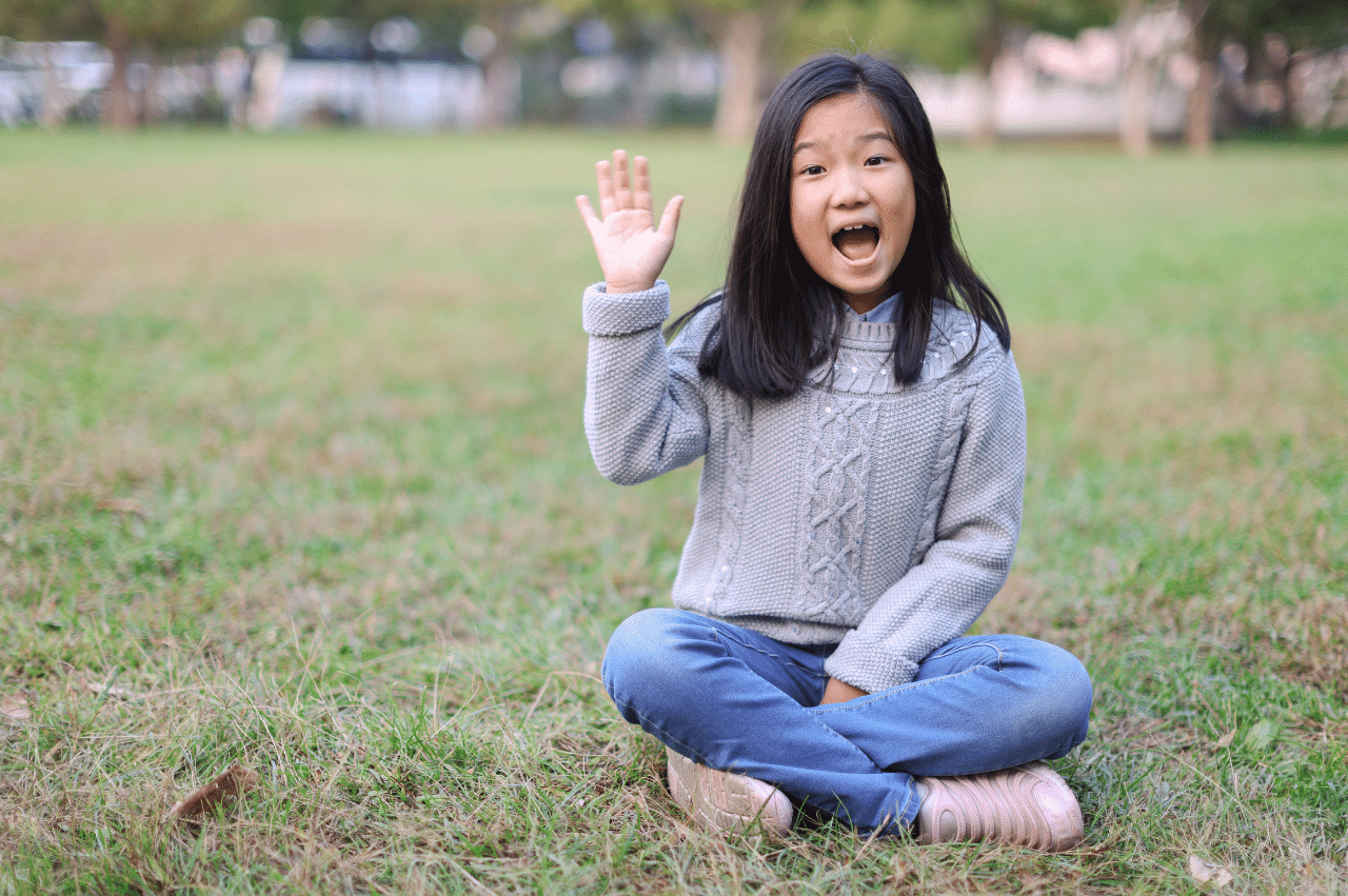 Photo of a young girl sitting in a patch of grass and waving