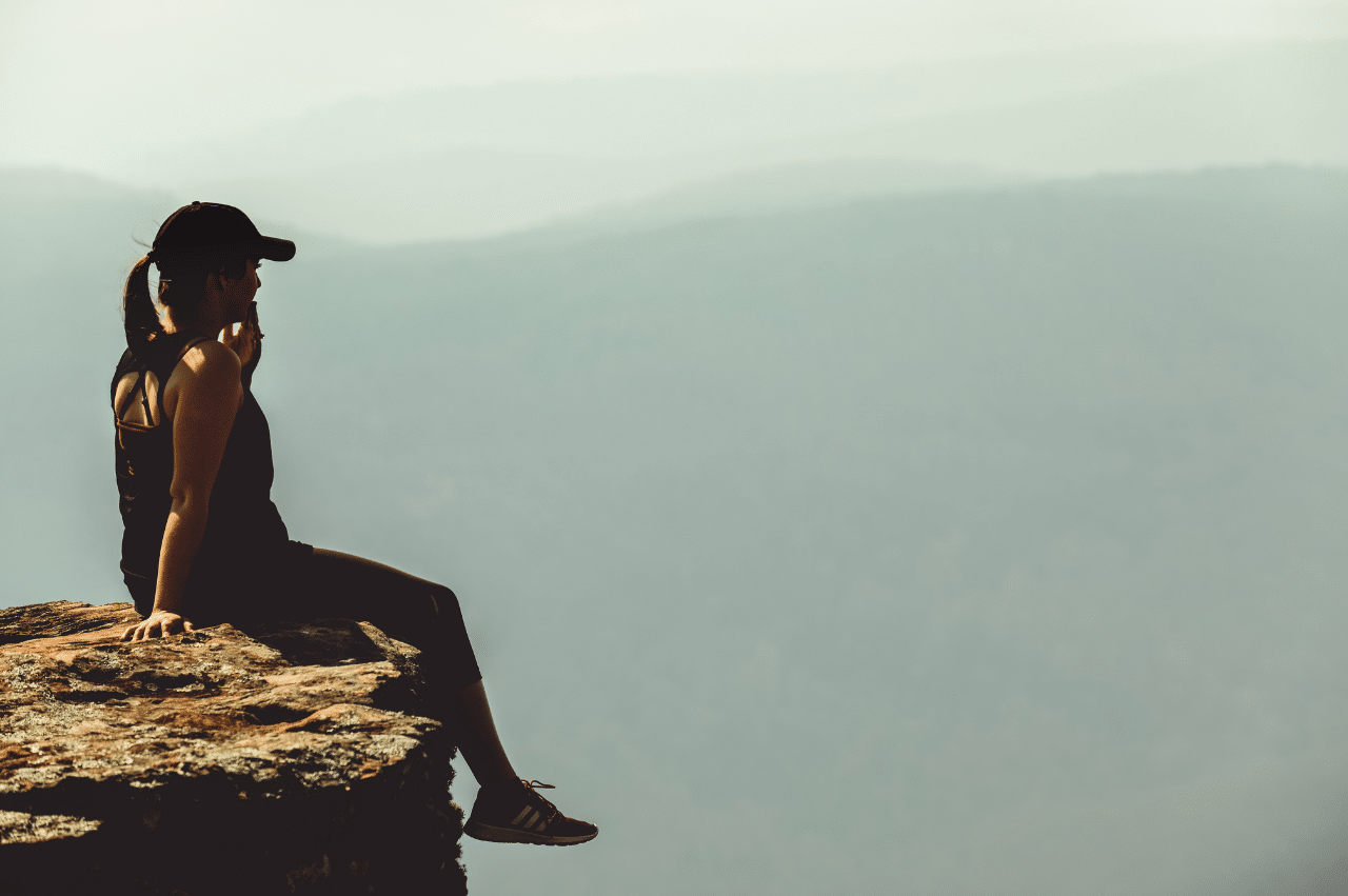 Picture of a woman sitting on a cliff. She is wearing a black cap and black workout clothes.