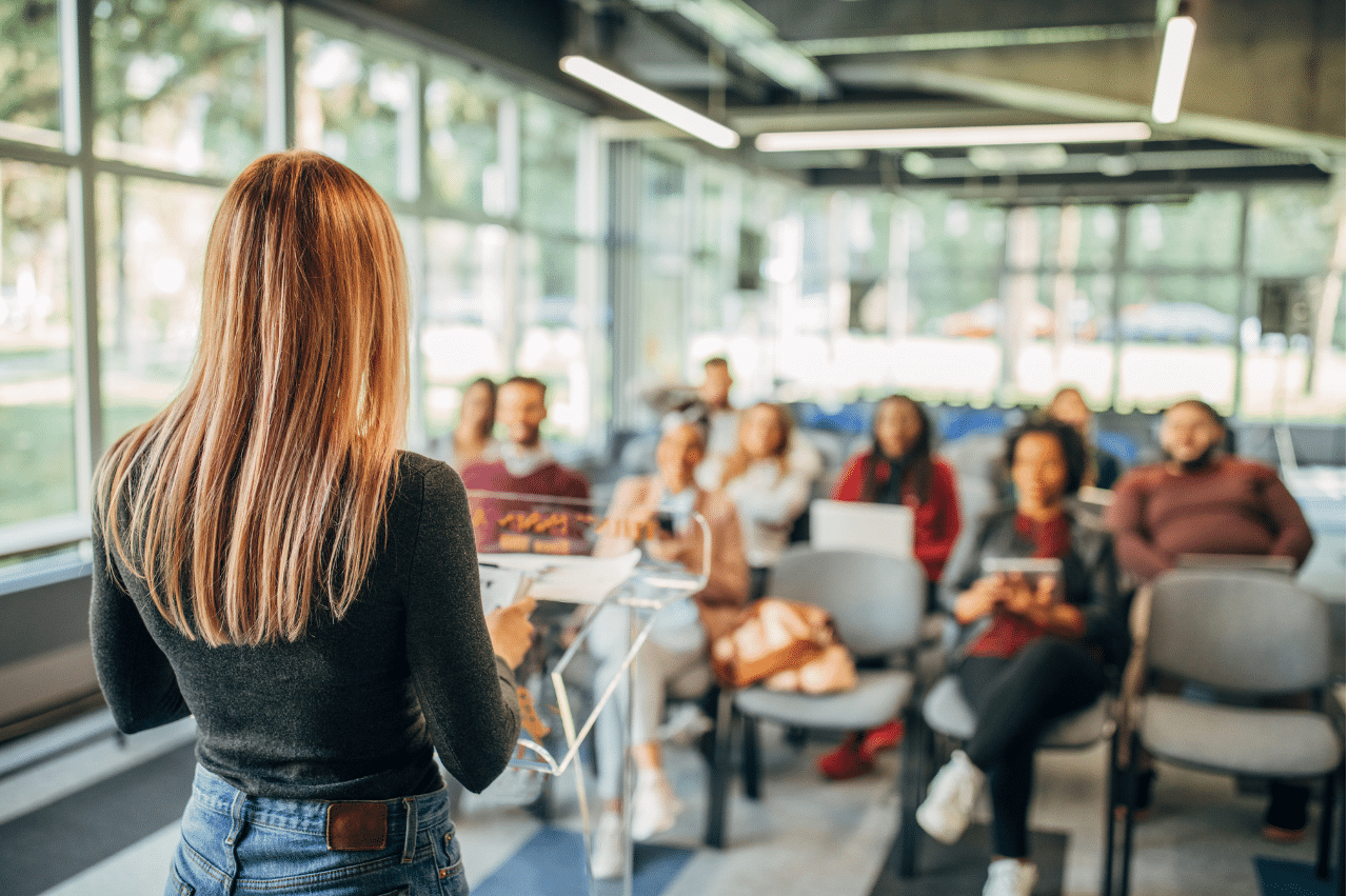 Photo of a woman standing in facing a small crowd of about 10 people sitting in gray chairs. The crowd is looking at her and are slightly blurred so the speaker is the focus.