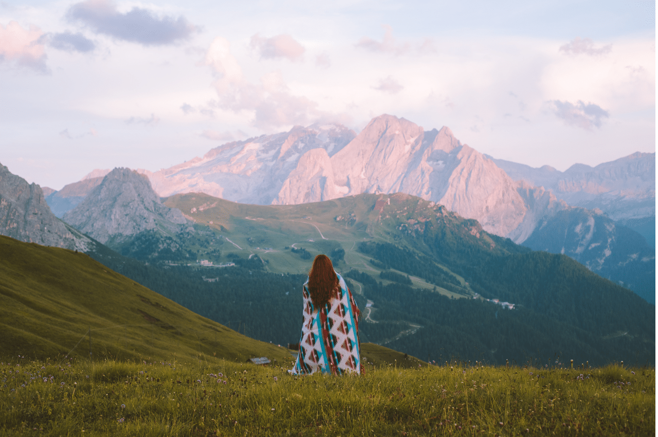 Photo of a mountain landscape. There is a woman standing with a blanket draped over her shoulders looking at the skyline.