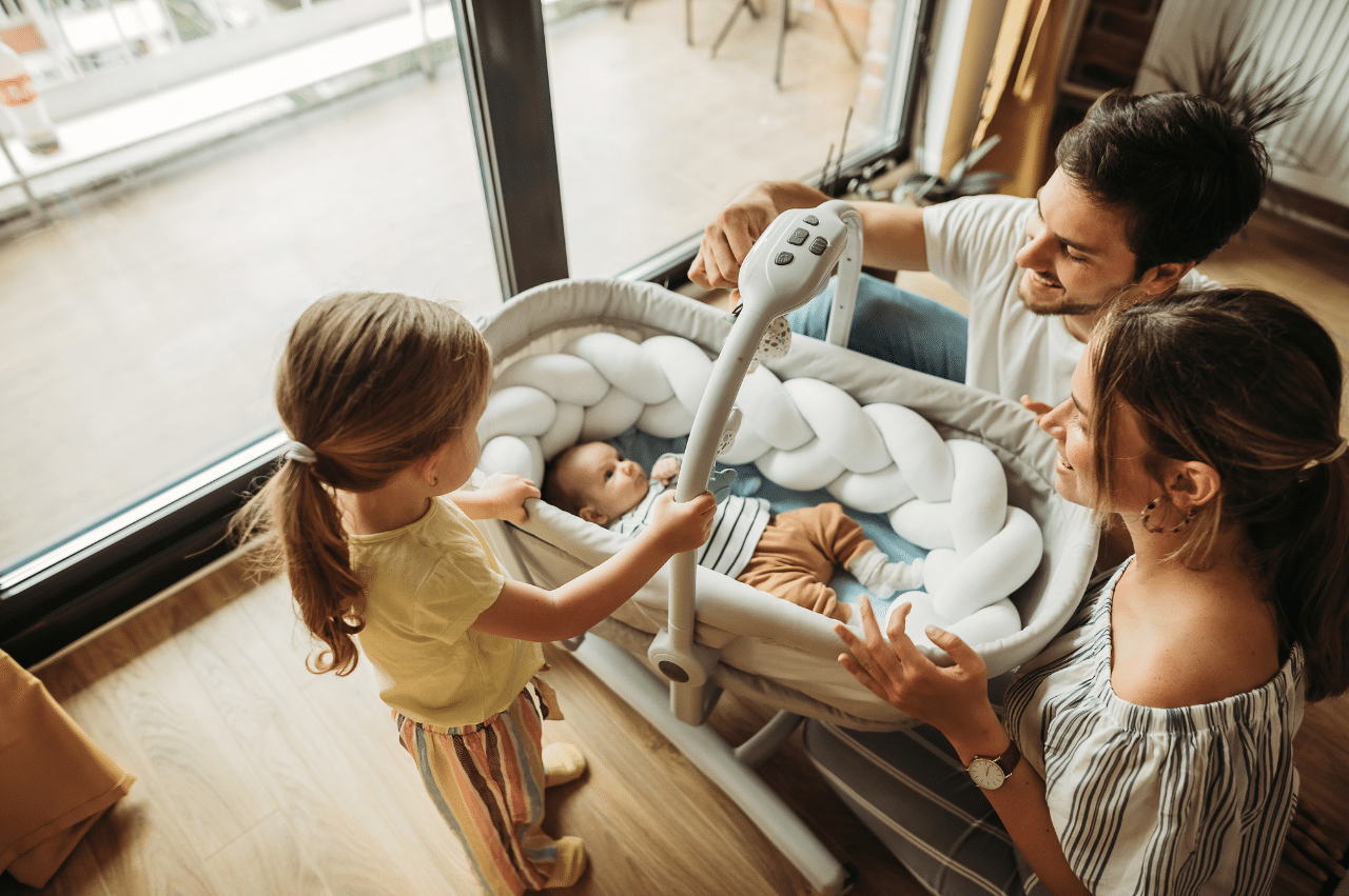 Photo of parents and a toddler older sister looking into a newborn baby's bassinet. The baby is looking at the dad who is smiling back.