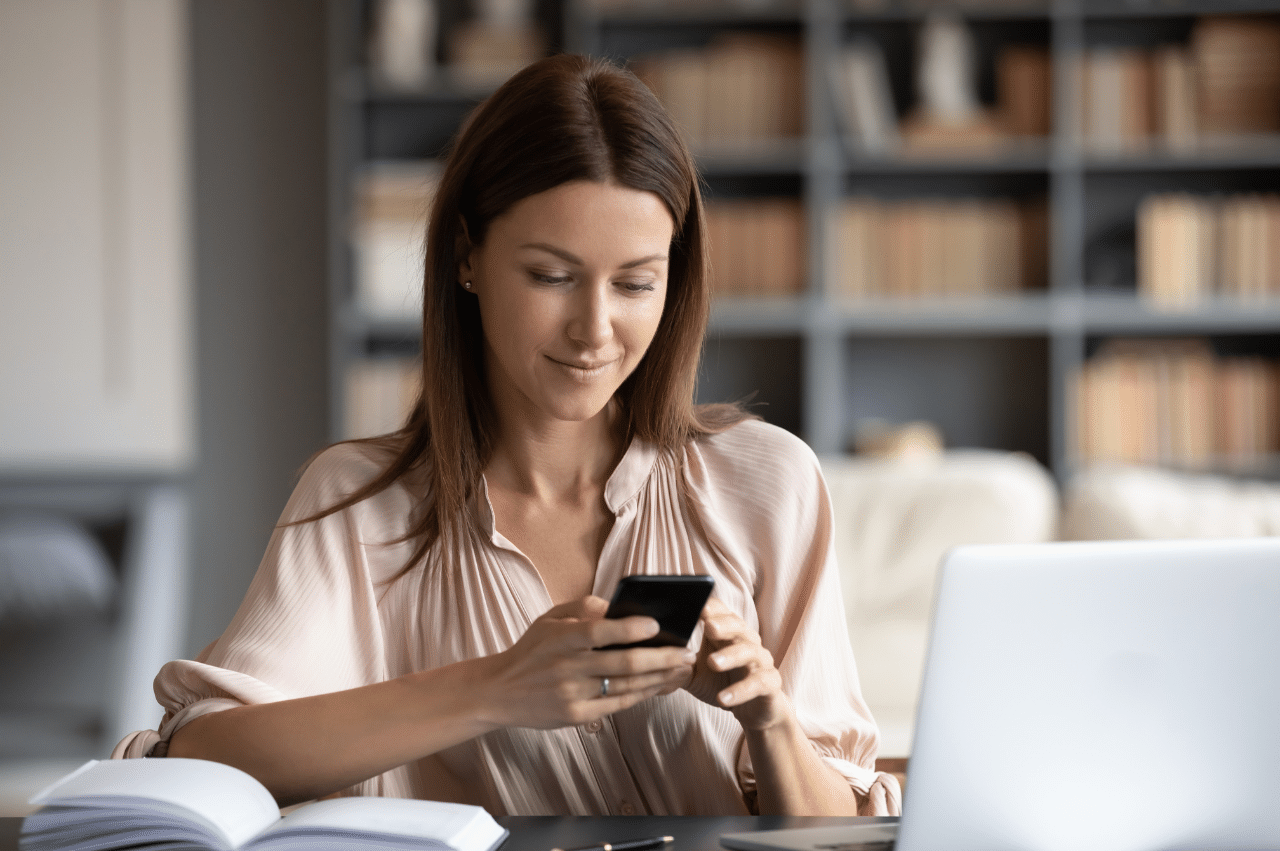 Photo of a woman looking at her phone sitting at a desk with a notebook and laptop in front of her.