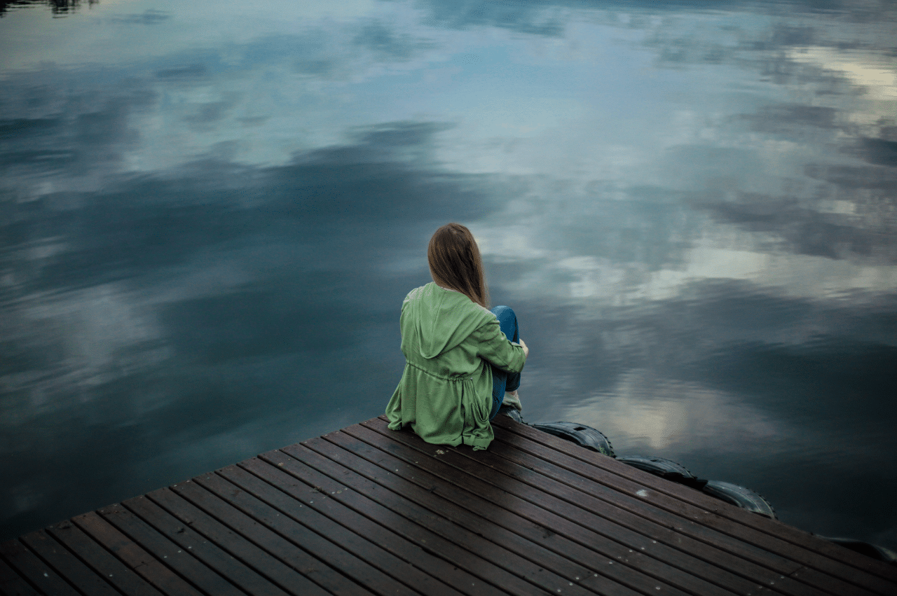 Photo of a woman sitting at the edge of a dock. The water is dark and blurry with the forms of clouds.