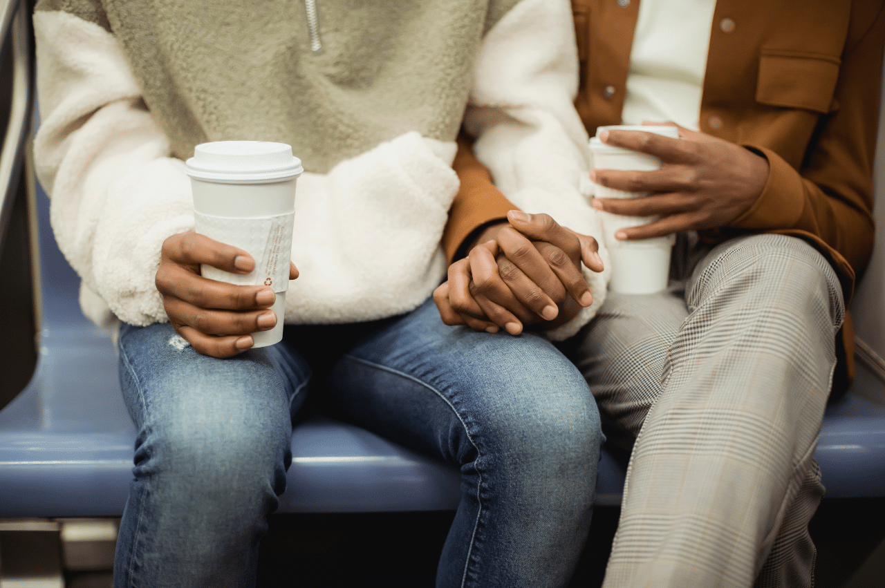 A photo of a Black couple holding hands. They are also holding hot drink cups and seem to be sitting on a subway train
