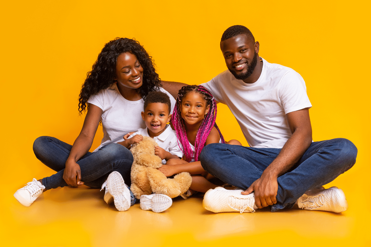Photo of a Black family sitting and smiling with a yellow backdrop. There are 2 parents and 2 children sitting between them.