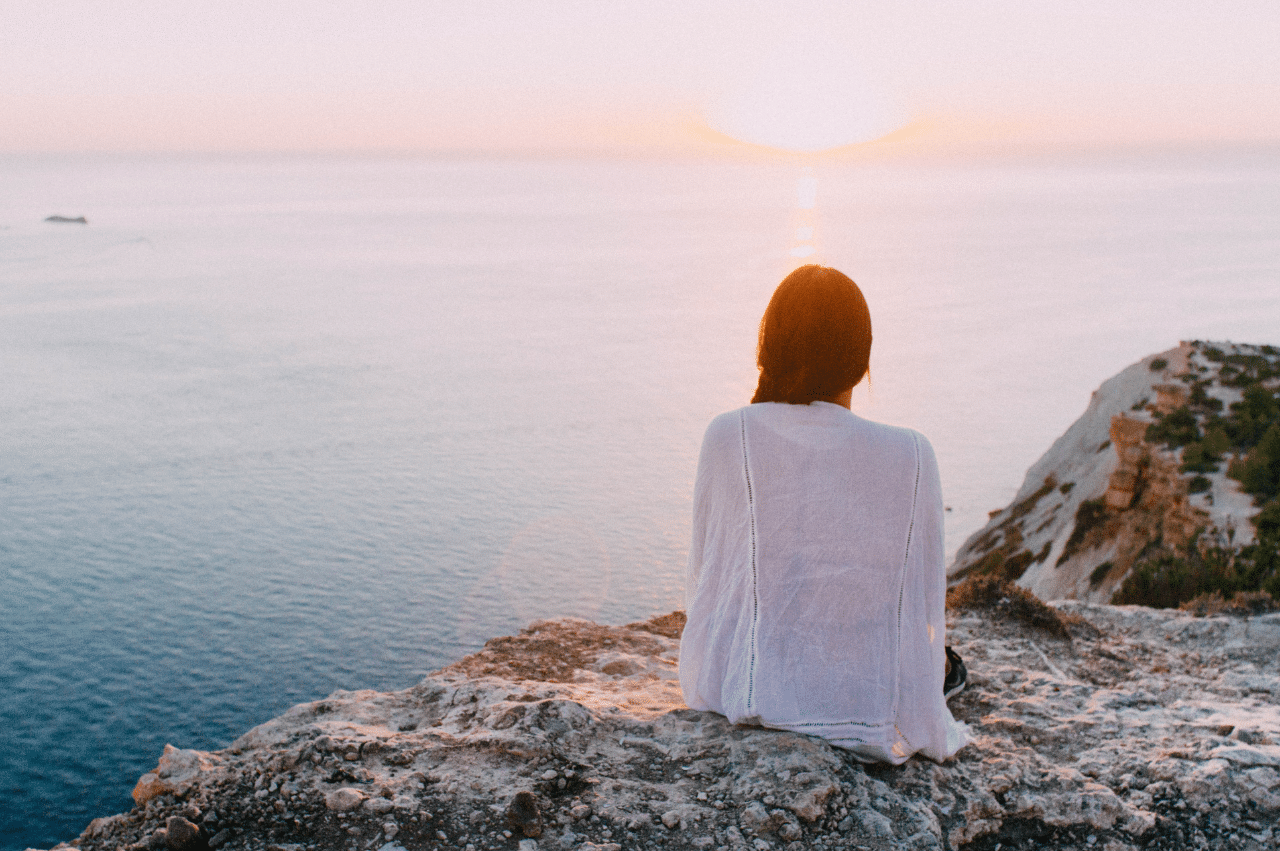 Photo of a woman sitting on a rock watching the sun rise/set over the ocean