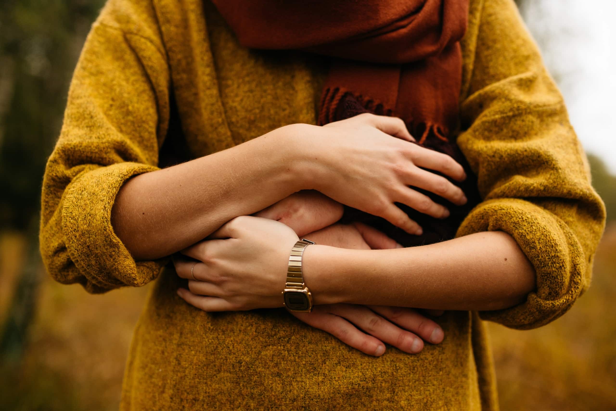 Woman wearing yellow sweater with her arms and a man's arms wrapped around her stomach.