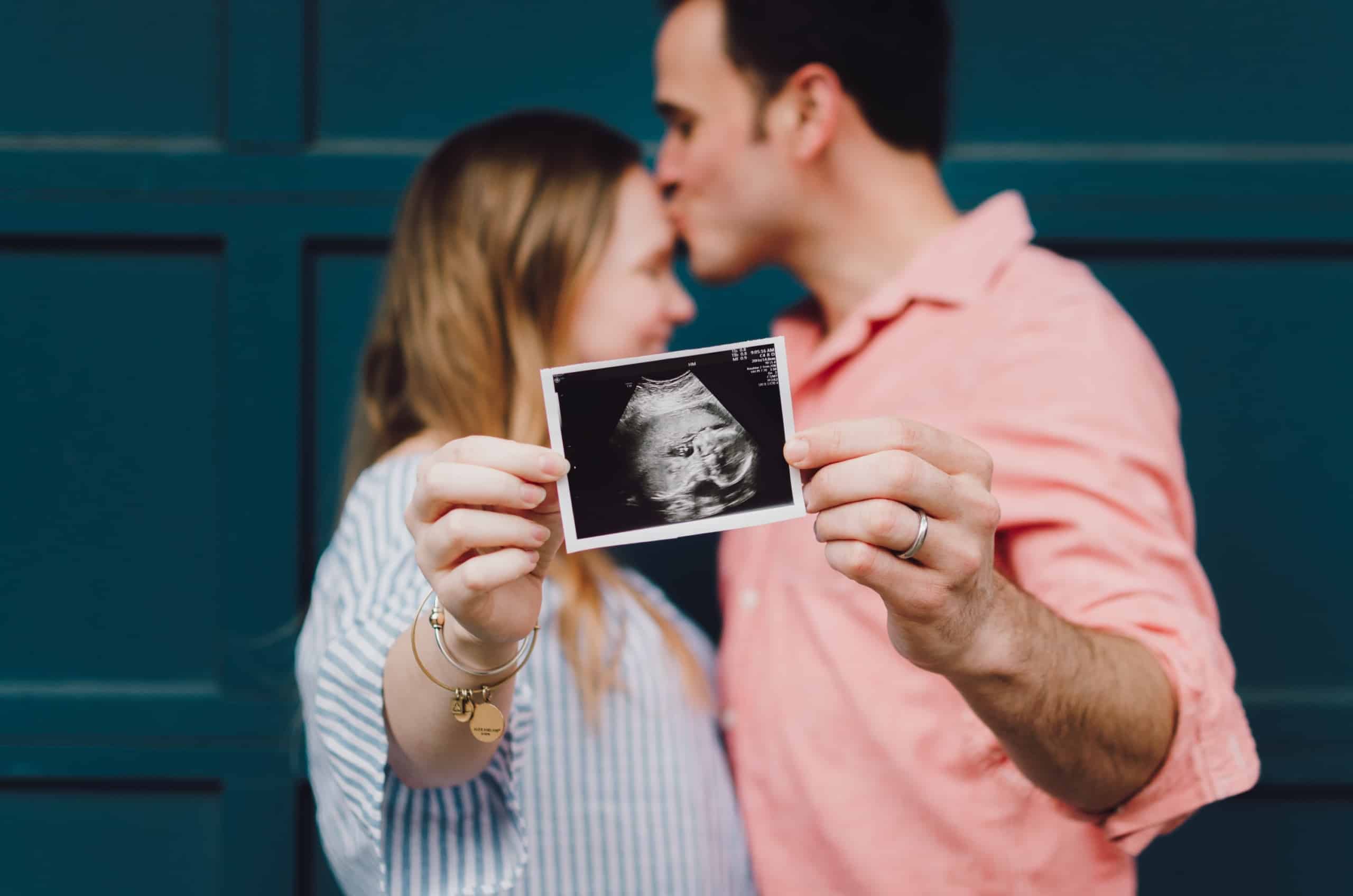 Man and woman leaning into one another and holding ultrasound photo up to camera.