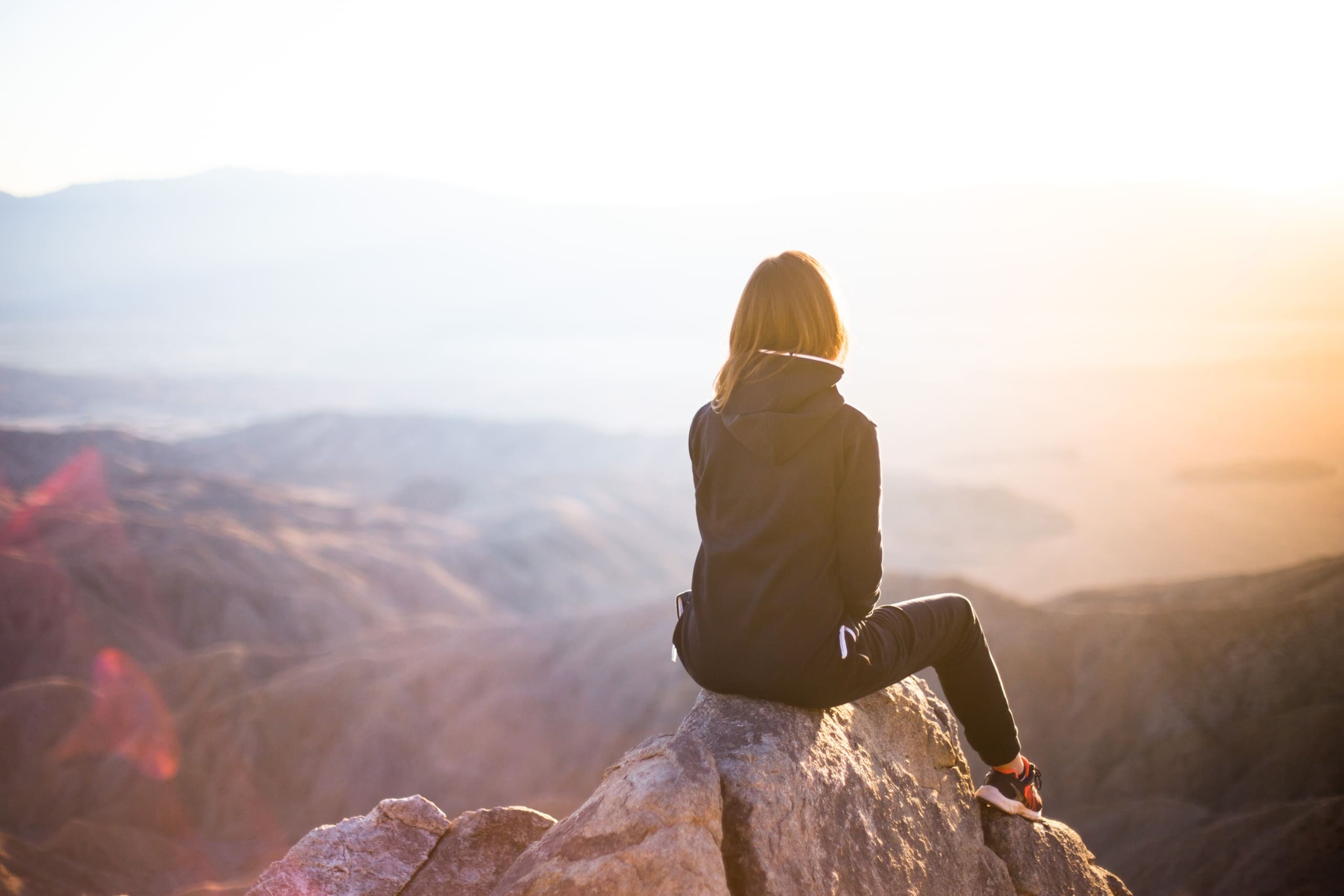 Woman with her back to the camera sitting on rocky mountain peak looking at sunrise.